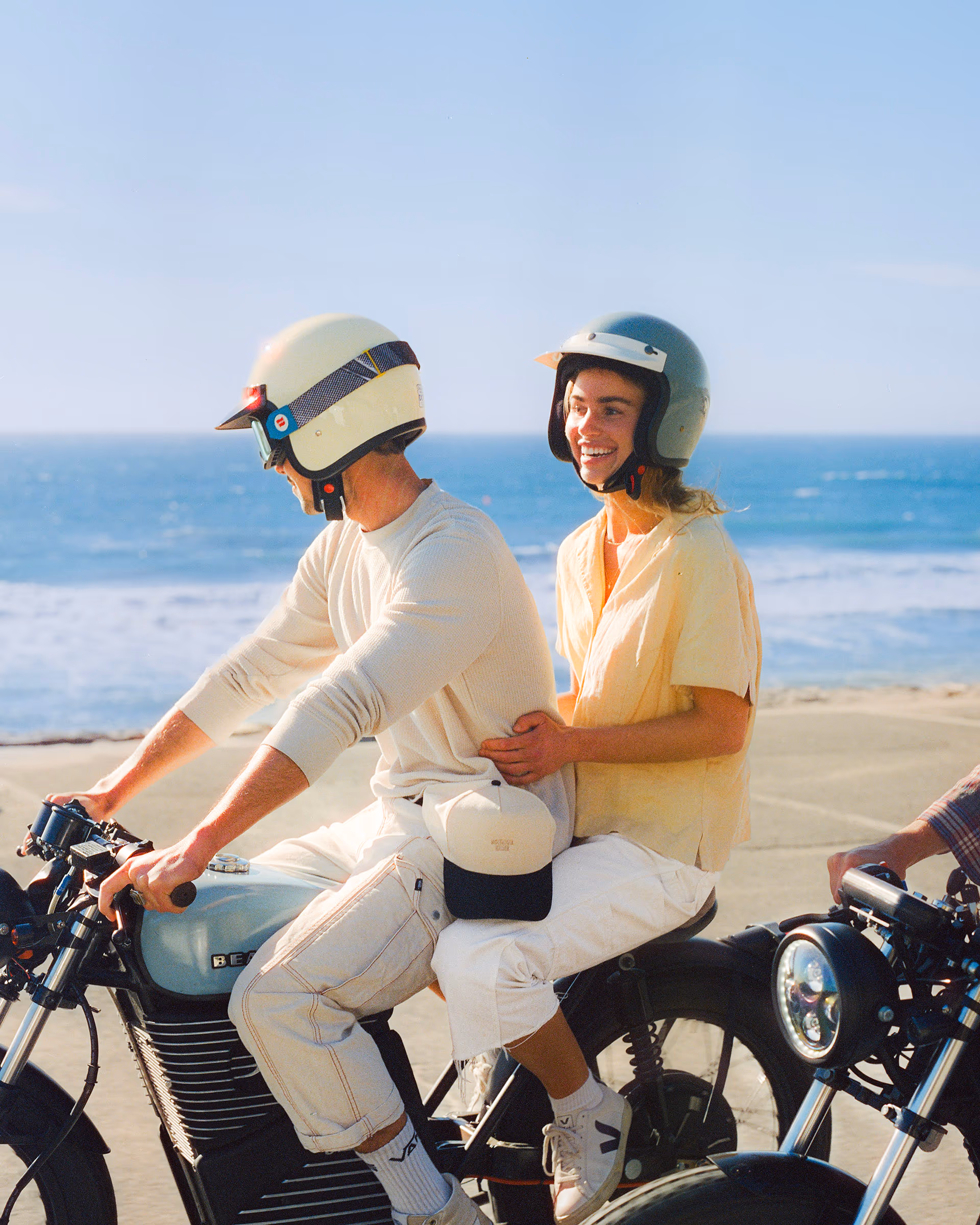 A couple riding two-up in matching cream helmets along the shore