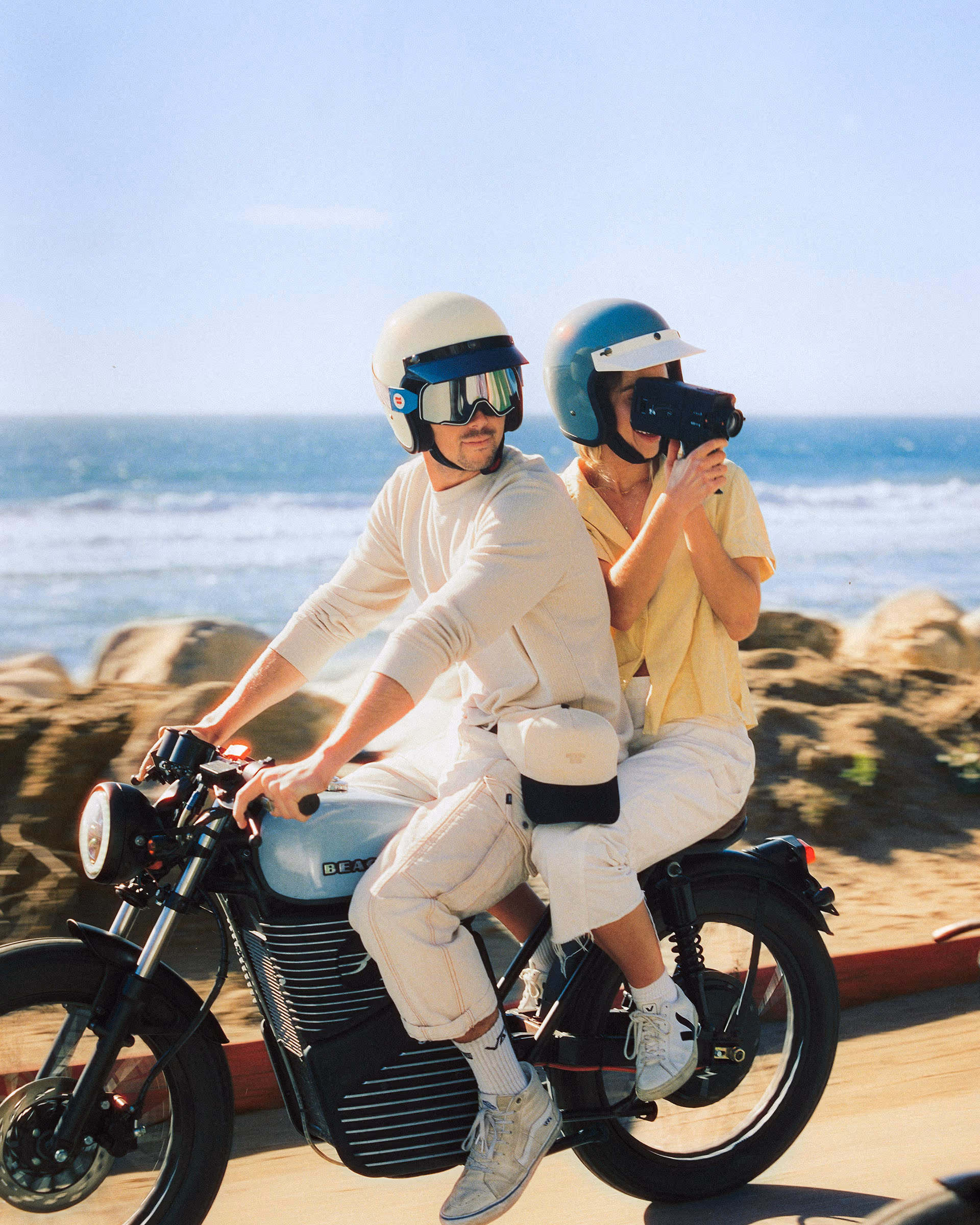 Two riders in cream helmets on a Beachman bike along the California coast, one filming on a camcorder