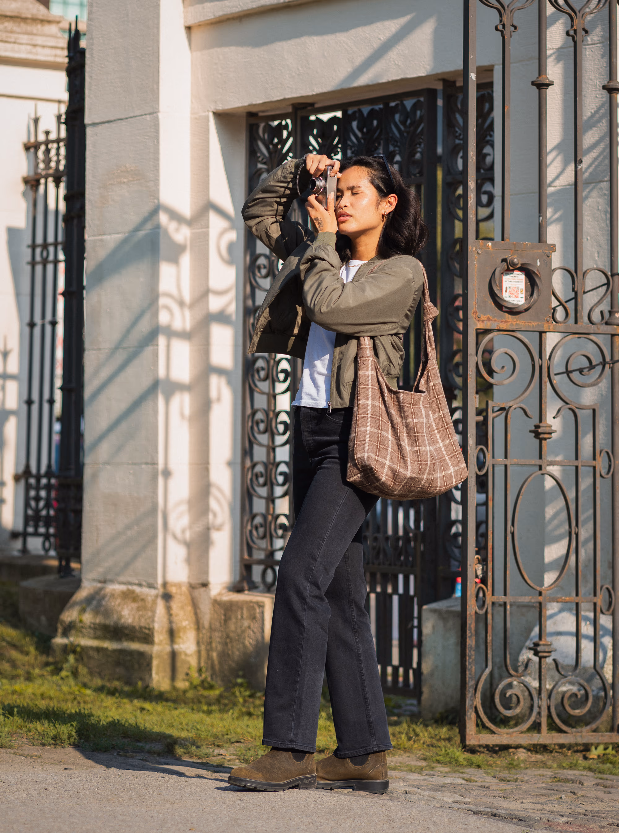 Shooting a film camera by a wrought-iron gate in olive Blundstones