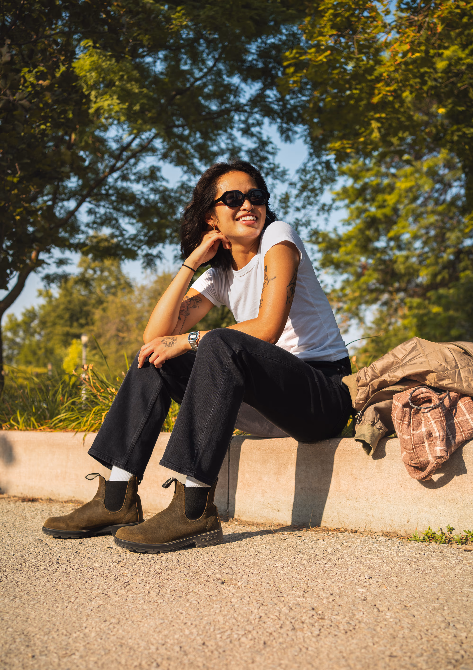 Sunglasses on, seated on a park ledge in the olive Chelsea boots