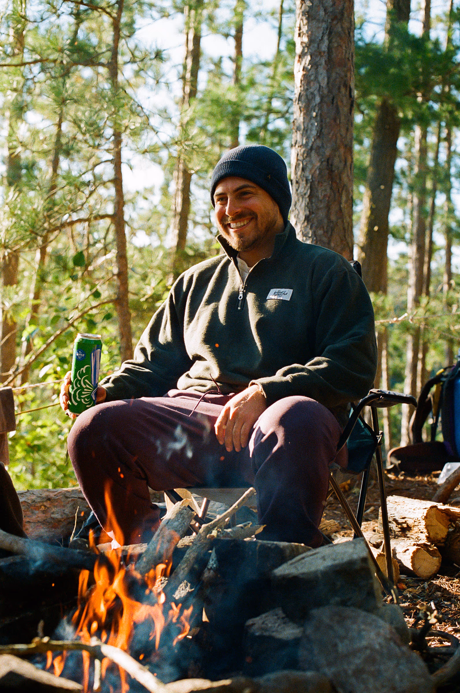 A camper in a beanie by the fire with a Steam Whistle pilsner, deep in the woods
