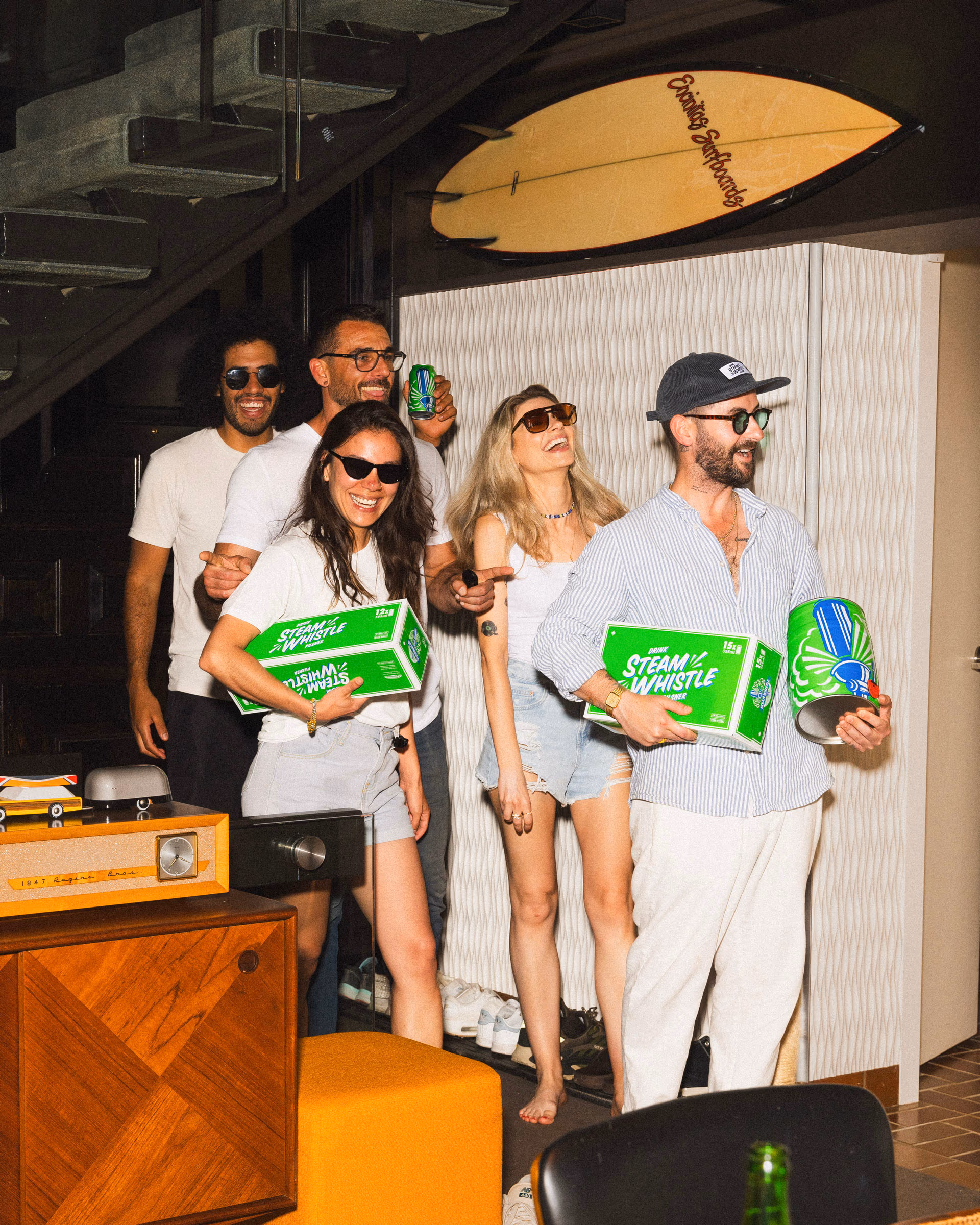 Friends holding Steam Whistle cases at a house party, surfboard on the wall