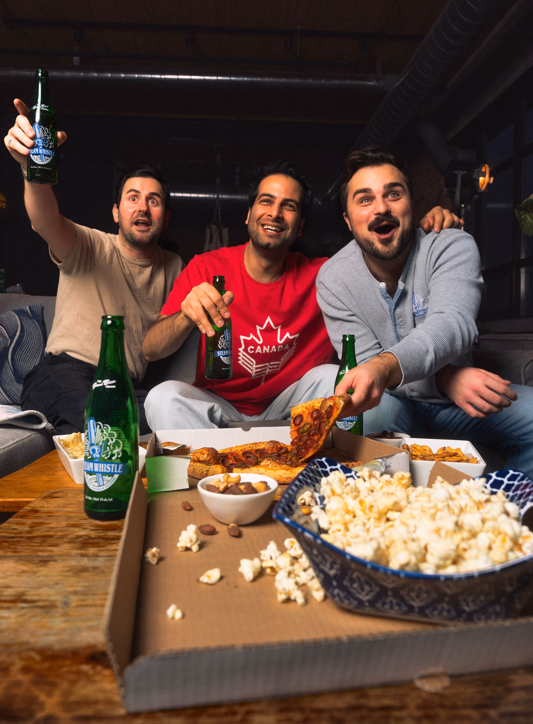 Three friends mid-cheer over pizza, popcorn and Steam Whistle on game night